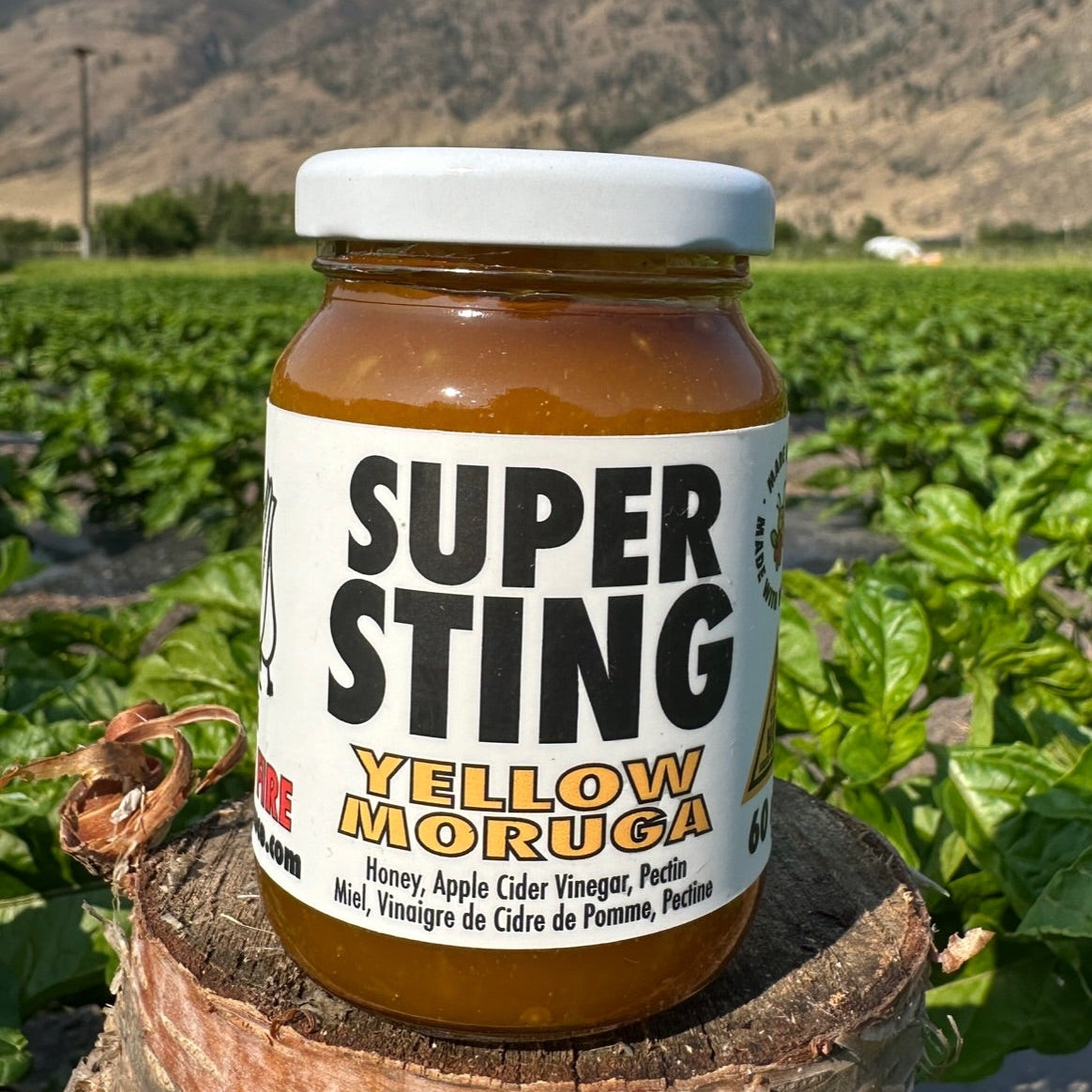 A jar of Super Sting Yellow Moruga hot sauce placed on a rustic wooden log in a lush green farm field with mountains in the background.